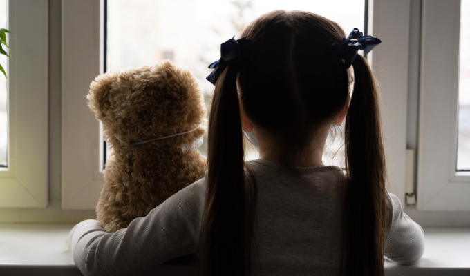 Menina triste com um ursinho olhando pela janela